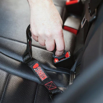 A close-up of a hand fastening a black car seat belt with red accents and a visible KONG brand tab, set against a dark textured car interior.