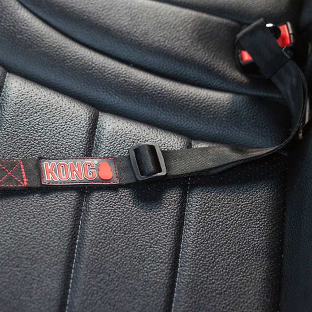 Close-up of a black car seat belt with a red and white KONG brand label and black plastic buckle, shown installed on a black textured leather seat.